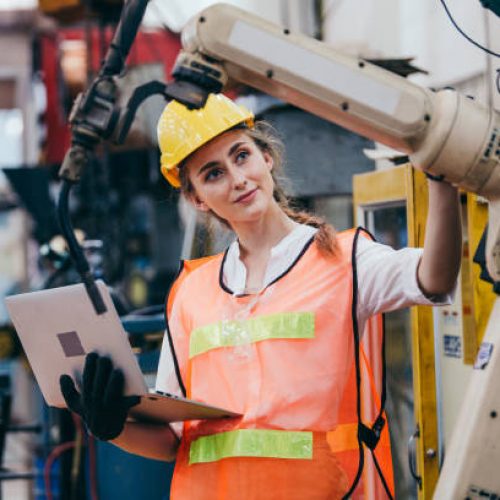 Female industrial engineer or technician worker in hard helmet and uniform using laptop checking on robotic arm machine. woman work hard in heavy technology invention industry manufacturing factory